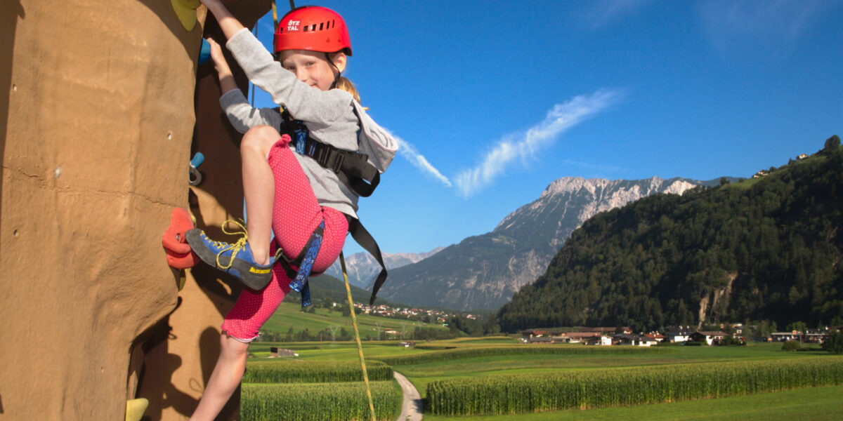 Ein kleines Kind, das einen Helm und einen Klettergurt trägt, klettert eine Felswand im Freien mit einer malerischen Berglandschaft im Hintergrund.