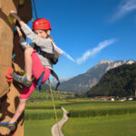 Ein kleines Kind, das einen Helm und einen Klettergurt trägt, klettert eine Felswand im Freien mit einer malerischen Berglandschaft im Hintergrund.