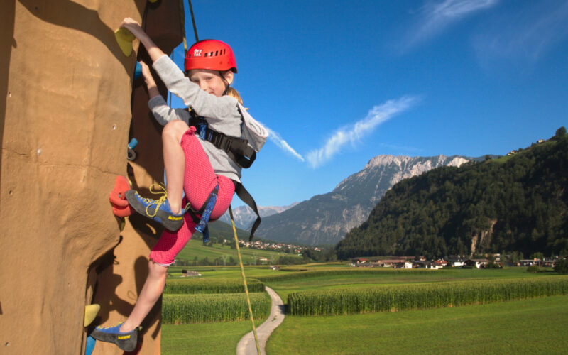 Ein kleines Kind, das einen Helm und einen Klettergurt trägt, klettert eine Felswand im Freien mit einer malerischen Berglandschaft im Hintergrund.