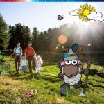 Eine vierköpfige Familie, die auf einer sonnenbeschienenen Wiese wandert, wobei das Bild von Cartoon-Schafen und Naturillustrationen überlagert wird.