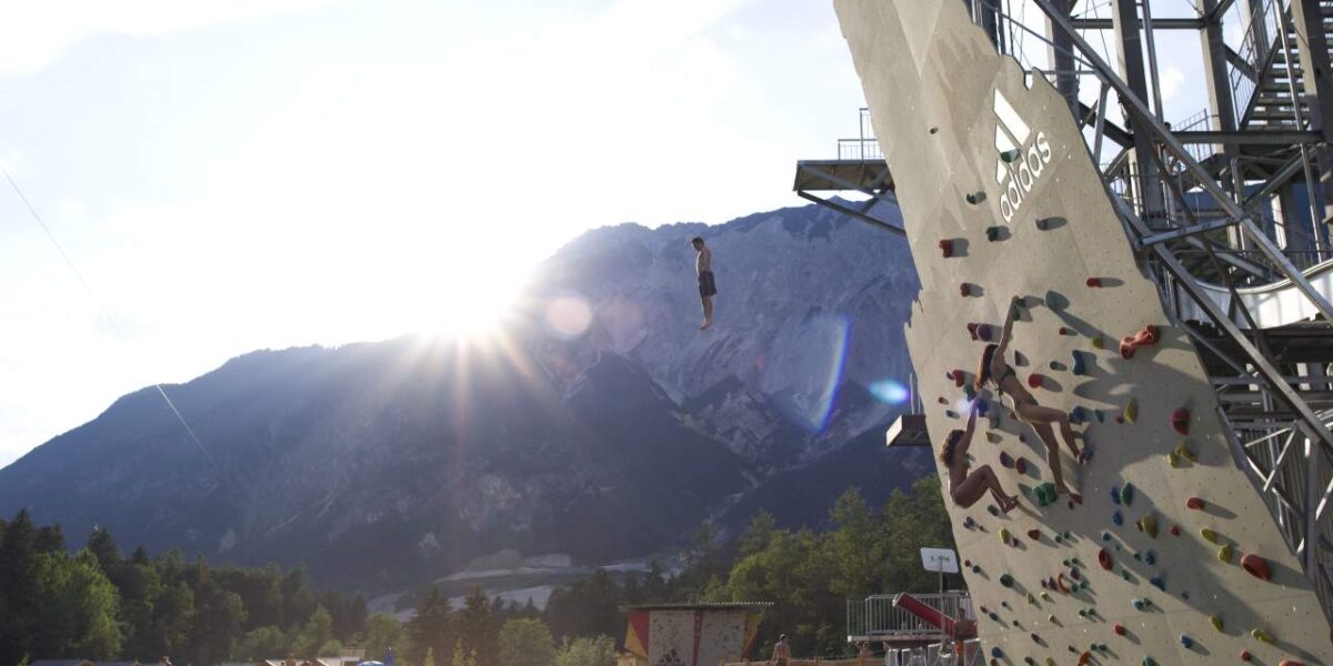 Eine Person springt von einer Kletterwand im Freien in einen See, mit Bergen und Bäumen im Hintergrund und der Sonne, die teilweise hinter den Gipfeln sichtbar ist.