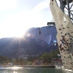 Eine Person springt von einer Kletterwand im Freien in einen See, mit Bergen und Bäumen im Hintergrund und der Sonne, die teilweise hinter den Gipfeln sichtbar ist.