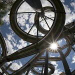 Ein Blick auf ineinander verschlungene Wasserrutschen aus Metall vor einem teilweise bewölkten Himmel, bei dem das Sonnenlicht durch die Rohre und Stützen scheint.