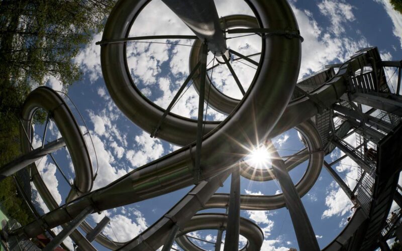 Ein Blick auf ineinander verschlungene Wasserrutschen aus Metall vor einem teilweise bewölkten Himmel, bei dem das Sonnenlicht durch die Rohre und Stützen scheint.