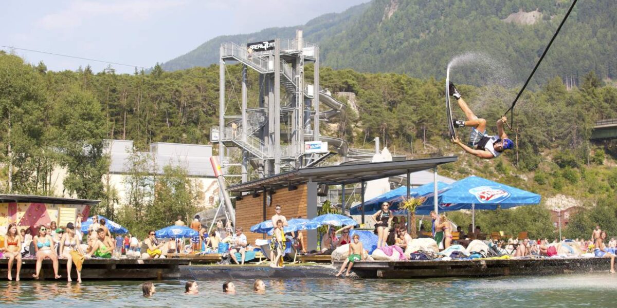 Eine Person schwingt sich an einer Seilrutsche in einen See, während die Leute zusehen und schwimmen; im Hintergrund sind eine Plattform am Wasser und Berge zu sehen.