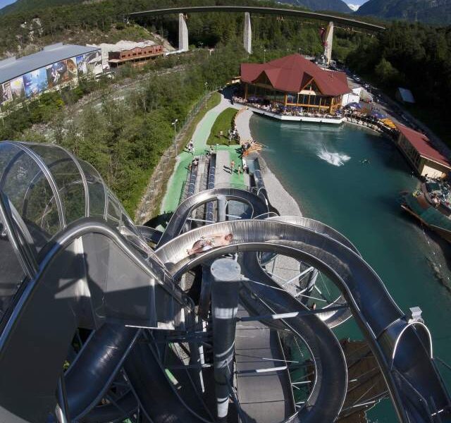 Ein Blick von der Spitze einer großen, kurvenreichen Wasserrutsche mit Blick auf einen Pool, Gebäude und eine Brücke mit Bergen im Hintergrund.