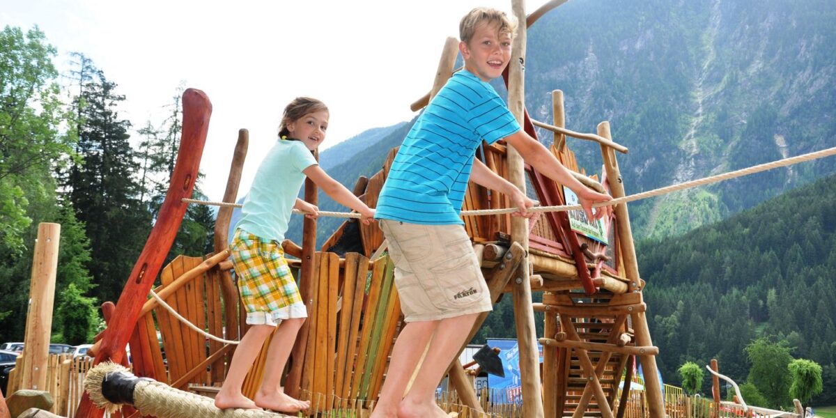 Zwei Kinder balancieren auf einer Seilbrücke auf einem Spielplatz im Freien mit Holzstrukturen, umgeben von Bäumen und Bergen.