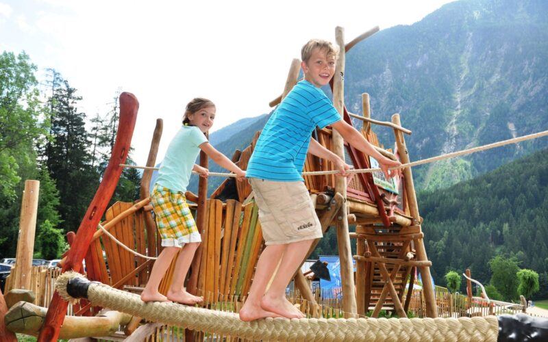 Zwei Kinder balancieren auf einer Seilbrücke auf einem Spielplatz im Freien mit Holzstrukturen, umgeben von Bäumen und Bergen.