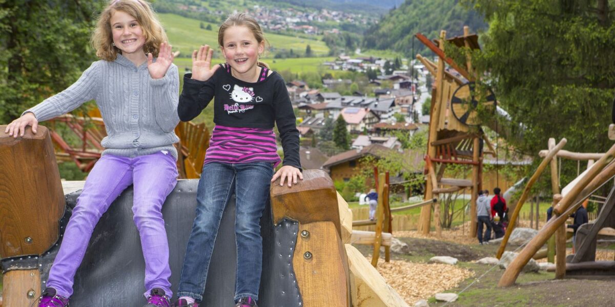 Zwei junge Mädchen sitzen auf einer Spielplatzrutsche im Freien, lächeln und winken in die Kamera, während im Hintergrund ein Dorf und Berge zu sehen sind.