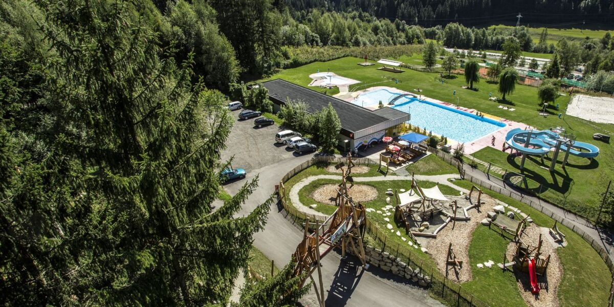 Luftaufnahme eines Freizeitgeländes mit Schwimmbad, Wasserrutsche, Spielplatz, Parkplatz und Bäumen in einer bergigen Landschaft.