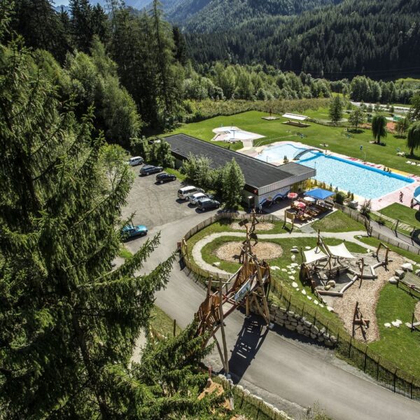 Luftaufnahme eines Freizeitgeländes mit Schwimmbad, Wasserrutsche, Spielplatz, Parkplatz und Bäumen in einer bergigen Landschaft.
