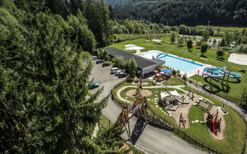 Luftaufnahme eines Freizeitgeländes mit Schwimmbad, Wasserrutsche, Spielplatz, Parkplatz und Bäumen in einer bergigen Landschaft.