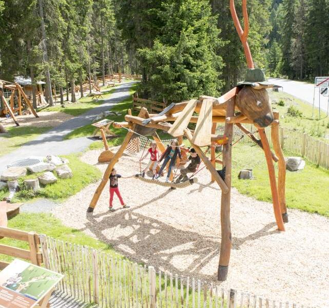Kinder spielen auf einem großen Holzspielplatz in Form eines Insekts, umgeben von Bäumen und einem eingezäunten Bereich in einem Park.
