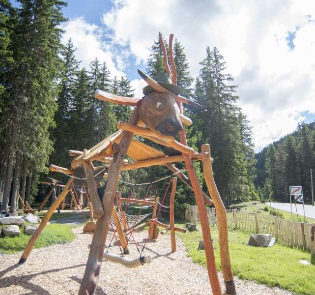 Spielplatz aus Holz in Form eines Rentiers, umgeben von Bäumen und auf einer Mulchfläche unter einem teilweise bewölkten Himmel.