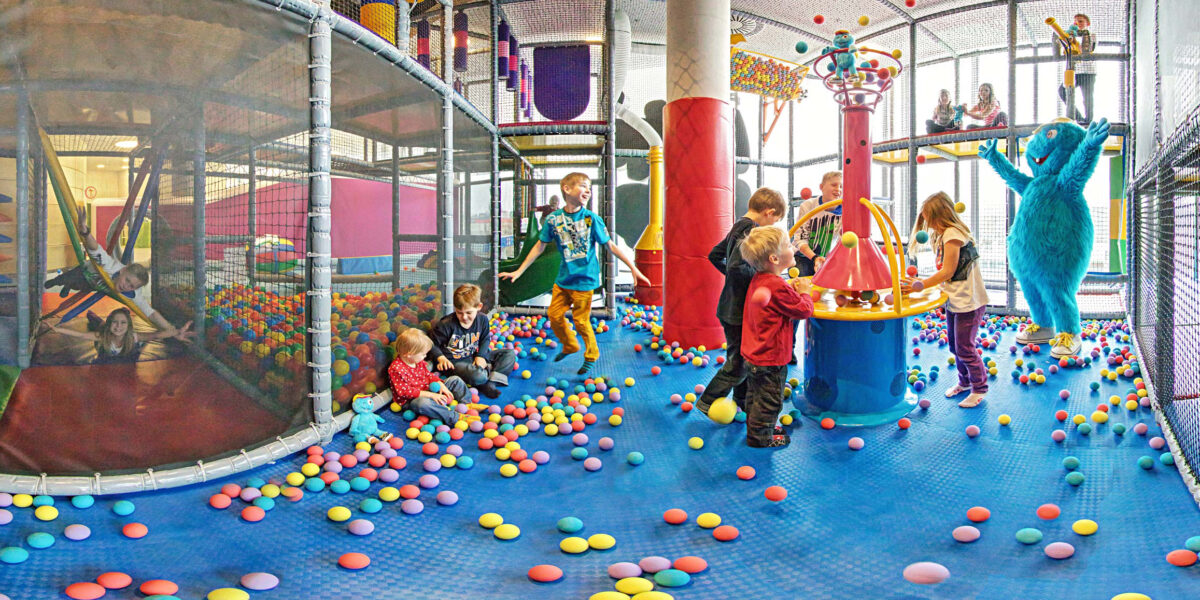 Kinder spielen auf einem Indoor-Spielplatz mit bunten Plastikbällen, Klettergerüsten und einer Person in einem blauen Maskottchenkostüm.