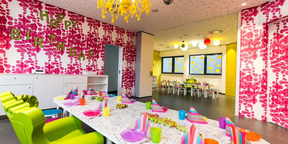 Ein bunt geschmückter Raum für eine Geburtstagsfeier, mit bunten Tellern, Tassen, Partyhüten und einem "Happy Birthday"-Schild an einer rosa gemusterten Wand.