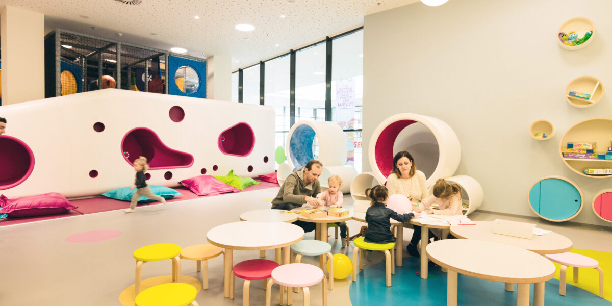 Kinder und Erwachsene sitzen an runden Tischen in einem farbenfrohen Indoor-Spielbereich mit weichen Sitzgelegenheiten, Wandregalen und einer großen Tunnelstruktur im Hintergrund.
