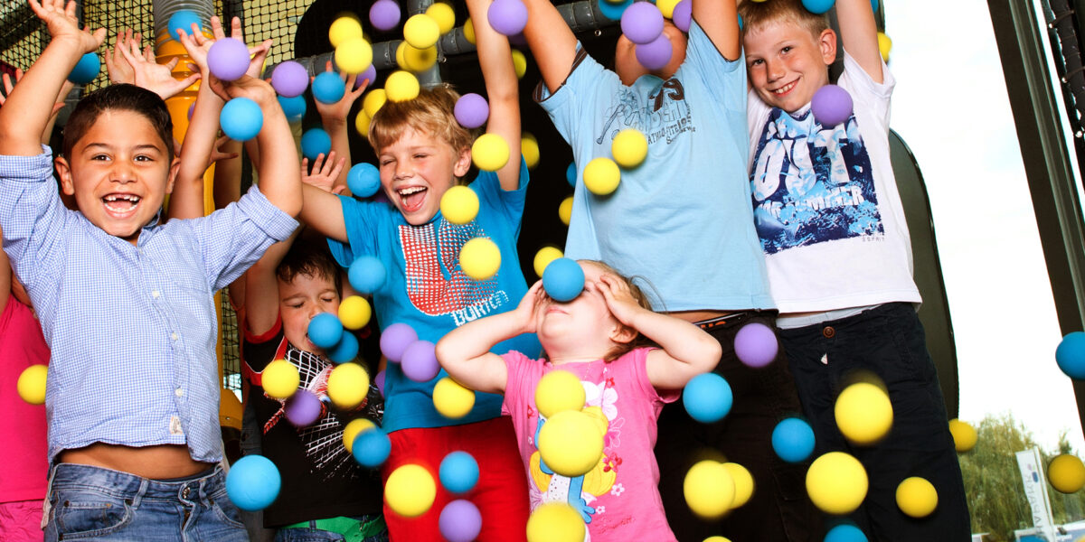 Eine Gruppe von Kindern steht mit erhobenen Armen und lächelnden Gesichtern unter herabfallenden bunten Plastikbällen in einem Indoor-Spielbereich.