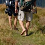 Drei Personen wandern durch eine Wiese, zwei mit Rucksäcken, eine barfuß mit Schuhen, die an ihrer Tasche hängen.