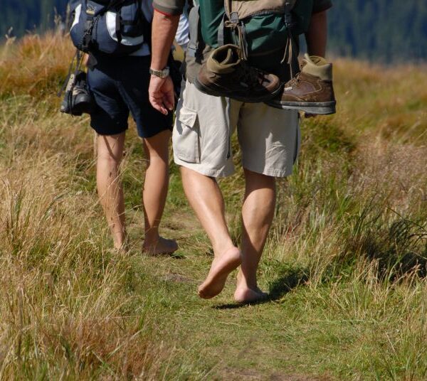 Drei Personen wandern durch eine Wiese, zwei mit Rucksäcken, eine barfuß mit Schuhen, die an ihrer Tasche hängen.