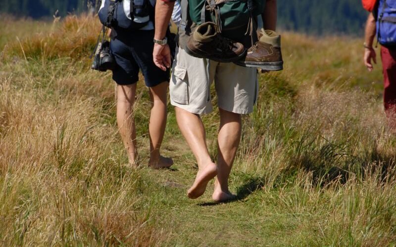 Drei Personen wandern durch eine Wiese, zwei mit Rucksäcken, eine barfuß mit Schuhen, die an ihrer Tasche hängen.