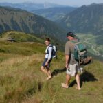 Zwei Personen mit Rucksäcken laufen barfuß über einen grasbewachsenen Bergpfad, mit einem malerischen Tal und Bergen im Hintergrund.