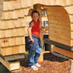 Ein Kind in rotem Hemd und blauer Hose steht an einem sonnigen Tag am Eingang eines Holzspielhauses mit Schindelverkleidung.