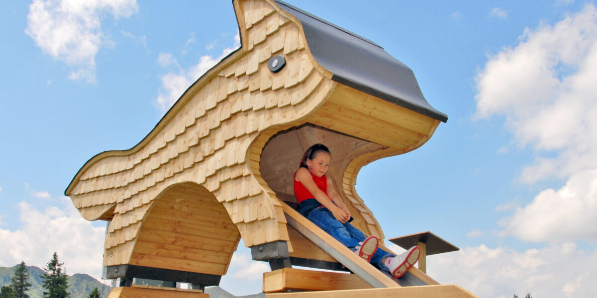 Ein Kind sitzt an einem sonnigen Tag im Freien in der Öffnung eines großen Spielplatzes aus Holz in Form eines Tieres, an dem eine Rutsche angebracht ist.