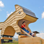 Ein Kind sitzt an einem sonnigen Tag im Freien in der Öffnung eines großen Spielplatzes aus Holz in Form eines Tieres, an dem eine Rutsche angebracht ist.