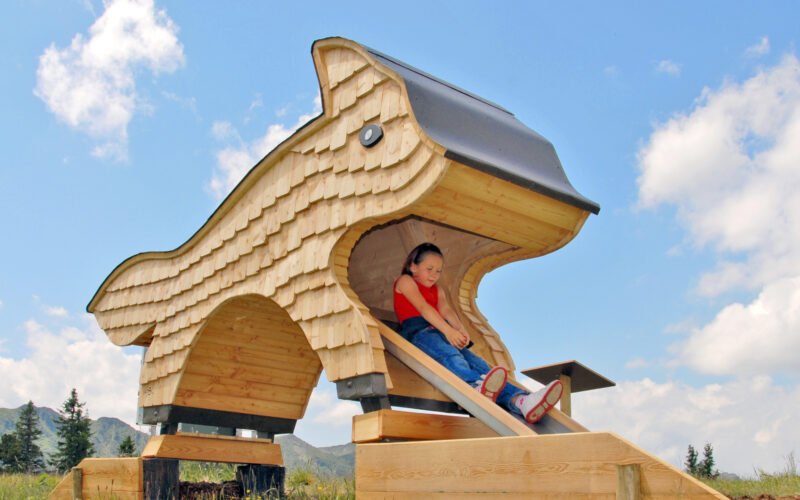 Ein Kind sitzt an einem sonnigen Tag im Freien in der Öffnung eines großen Spielplatzes aus Holz in Form eines Tieres, an dem eine Rutsche angebracht ist.