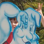 Ein Kind schaut durch den Ausschnitt einer Zeichentrickfigur, die Eiscreme isst, während ein anderes Kind hinter dem Ausschnitt spielerisch die Geste „Hasenohren“ macht.