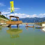 Holzplattformen und eine kleine überdachte Struktur stehen in einem flachen grünlichen Teich, umgeben von grasbewachsenen Hügeln und Bergen unter einem klaren blauen Himmel.