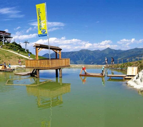 Holzplattformen und eine kleine überdachte Struktur stehen in einem flachen grünlichen Teich, umgeben von grasbewachsenen Hügeln und Bergen unter einem klaren blauen Himmel.