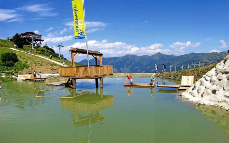 Holzplattformen und eine kleine überdachte Struktur stehen in einem flachen grünlichen Teich, umgeben von grasbewachsenen Hügeln und Bergen unter einem klaren blauen Himmel.