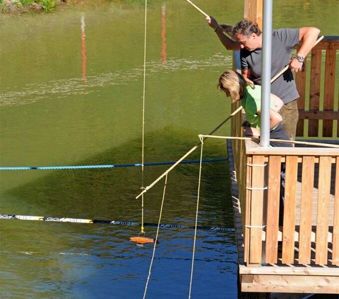 Zwei Personen stehen auf einem Holzdeck über einem Teich und angeln mit langen Bambusstangen, an deren Schnüren flache Gegenstände befestigt sind, die das Wasser berühren.