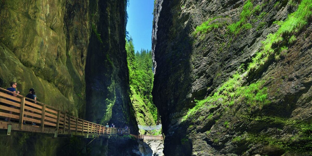 Eine schmale Schlucht mit steilen Felsen, grüner Vegetation und einem türkisfarbenen Fluss; man geht auf einem Holzpfad, der an der Felswand befestigt ist.
