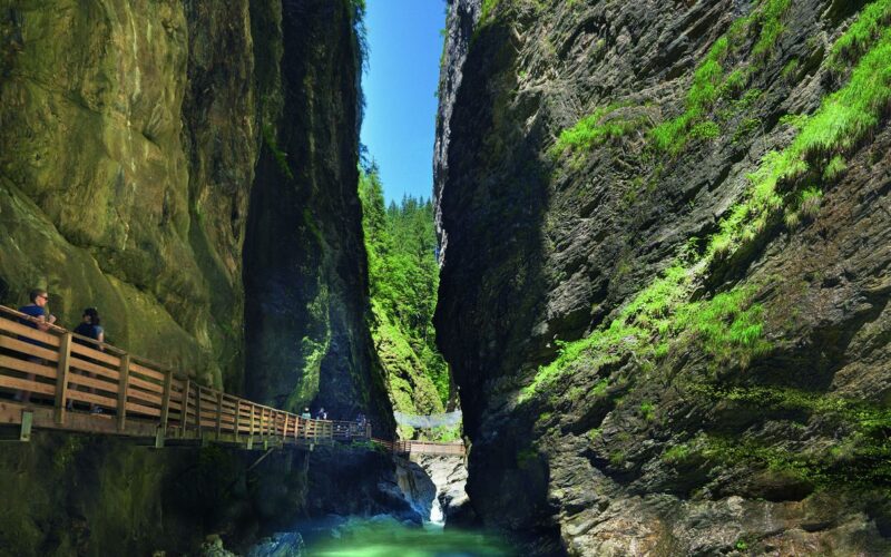 Eine schmale Schlucht mit steilen Felsen, grüner Vegetation und einem türkisfarbenen Fluss; man geht auf einem Holzpfad, der an der Felswand befestigt ist.