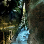 Eine enge, felsige Schlucht mit einem rauschenden Bach und einem Holzsteg auf der linken Seite, der von oben beleuchtet wird.