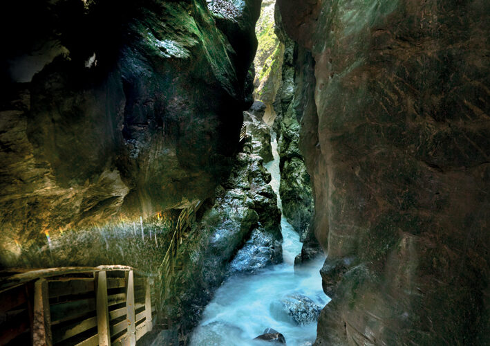 Eine enge, felsige Schlucht mit einem rauschenden Bach und einem Holzsteg auf der linken Seite, der von oben beleuchtet wird.