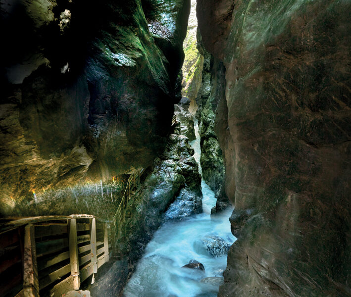 Eine enge, felsige Schlucht mit einem rauschenden Bach und einem Holzsteg auf der linken Seite, der von oben beleuchtet wird.