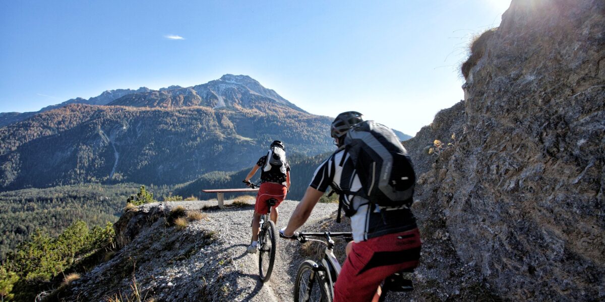 Zwei Radfahrer mit Helmen und Rucksäcken fahren mit Mountainbikes auf einem schmalen Schotterweg mit einem Berg und blauem Himmel im Hintergrund.