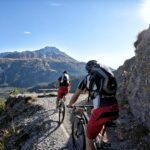 Zwei Radfahrer mit Helmen und Rucksäcken fahren mit Mountainbikes auf einem schmalen Schotterweg mit einem Berg und blauem Himmel im Hintergrund.