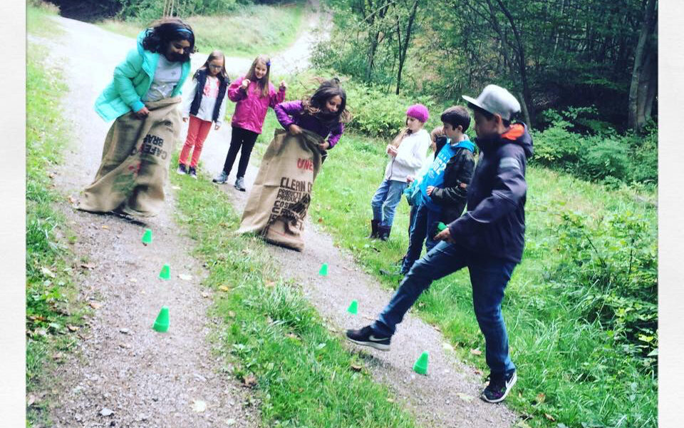 Kinder nehmen an einem Sackhüpfen auf einem Schotterweg im Freien teil, während ein Mann die Aufsicht führt; grüne Kegel markieren die von Bäumen umgebene Strecke.