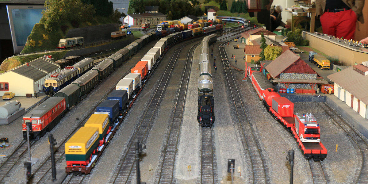 Modelleisenbahnanlage mit mehreren Gleisen, Güterzügen, Gebäuden, Fahrzeugen und Miniaturfiguren, die in einer Halle aufgebaut ist, während die Menschen im Hintergrund zusehen.
