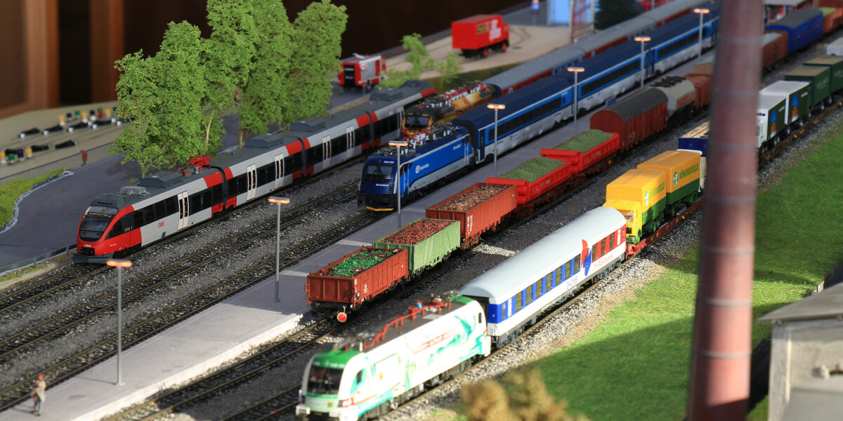 Eine detaillierte Modelleisenbahnanlage mit verschiedenen bunten Lokomotiven und Güterwagen auf mehreren Gleisen, mit Miniaturbäumen und Gebäuden im Hintergrund.