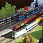 Eine detaillierte Modelleisenbahnanlage mit verschiedenen bunten Lokomotiven und Güterwagen auf mehreren Gleisen, mit Miniaturbäumen und Gebäuden im Hintergrund.