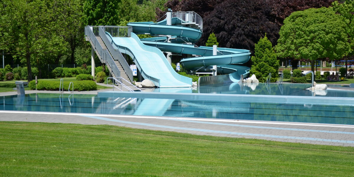 Ein leeres Außenschwimmbecken mit einer großen, hellblauen Spiralrutsche ist von grünem Gras und Bäumen umgeben.