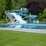 Ein leeres Außenschwimmbecken mit einer großen, hellblauen Spiralrutsche ist von grünem Gras und Bäumen umgeben.