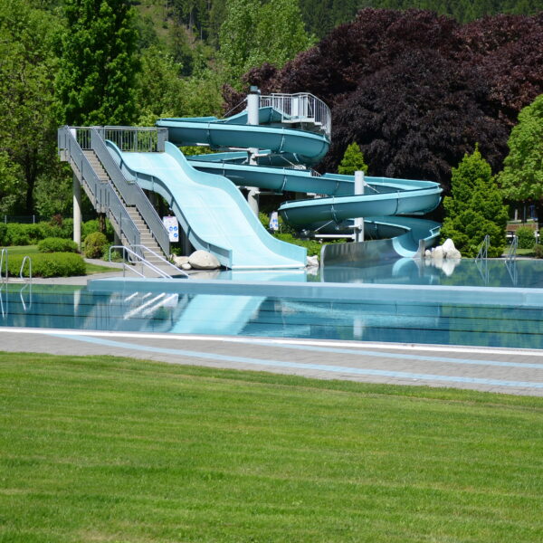 Ein leeres Außenschwimmbecken mit einer großen, hellblauen Spiralrutsche ist von grünem Gras und Bäumen umgeben.