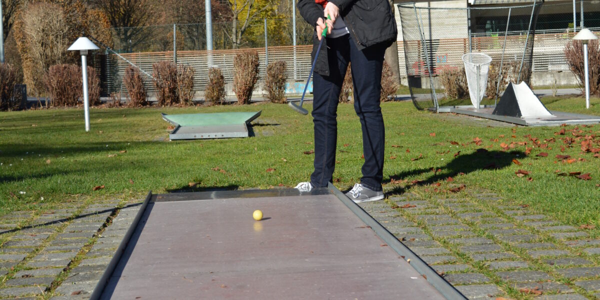 Eine Person, die im Freien Minigolf spielt, bereitet sich darauf vor, einen gelben Golfball auf einem schmalen Platz mit Bäumen und Hügeln im Hintergrund zu schlagen.